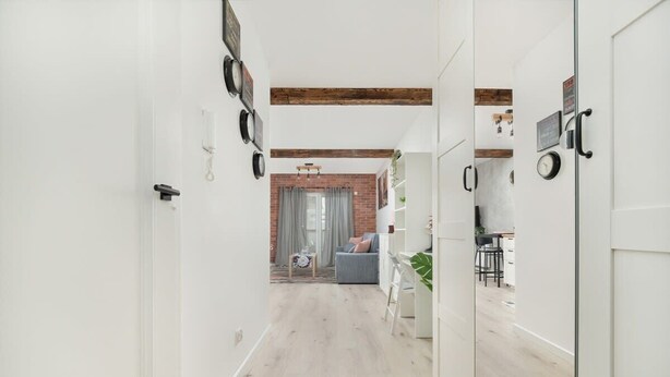 Ein Flur mit hellem Holzboden, weißen Wänden und freiliegenden Backsteinen im Hintergrund. Es gibt eine Sitzecke mit einem grauen Sofa und einem kleinen Tisch mit einer Pflanze.