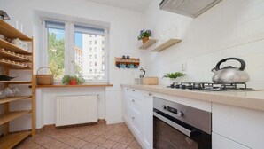 Una cocina compacta con armarios blancos, estufa a gas y horno integrado. Hay estantes con utensilios de cocina y una ventana con vistas a edificios.