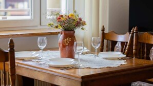 Una mesa de comedor de madera dispuesta con platos blancos, cubiertos y copas de vino, junto con un florero con flores.