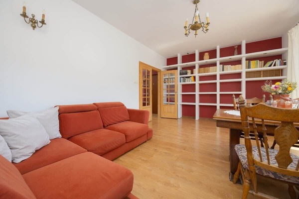 A spacious living room with light wooden floors, red armchairs, and table. The large bookshelf in the background adds warmth to the room.