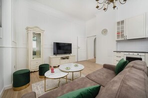 Un salon moderne doté d’un téléviseur à écran plat, de deux petites tables rondes, d’un canapé et d’un coin cuisine avec des placards et une horloge.