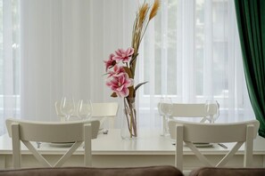Una mesa de comedor con sillas blancas, un florero con flores rosadas y una ventana con cortinas translúcidas.