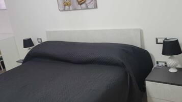 Un lit bien fait, recouvert d’une couverture foncée, une table de chevet avec une lampe et une œuvre d’art accrochée au mur.