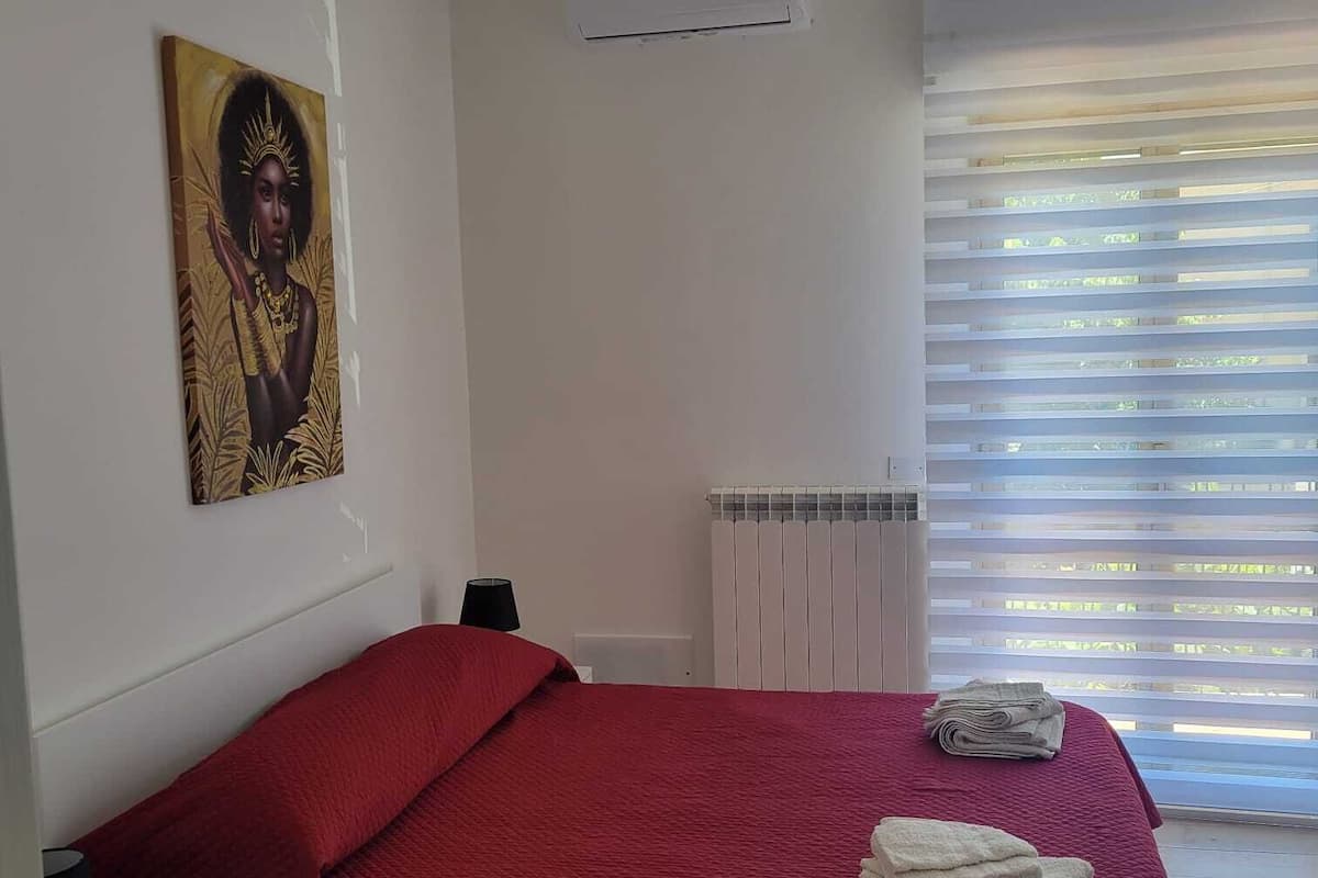 A bedroom with a red bedspread, a radiator, an air conditioning unit, and a window with blinds.