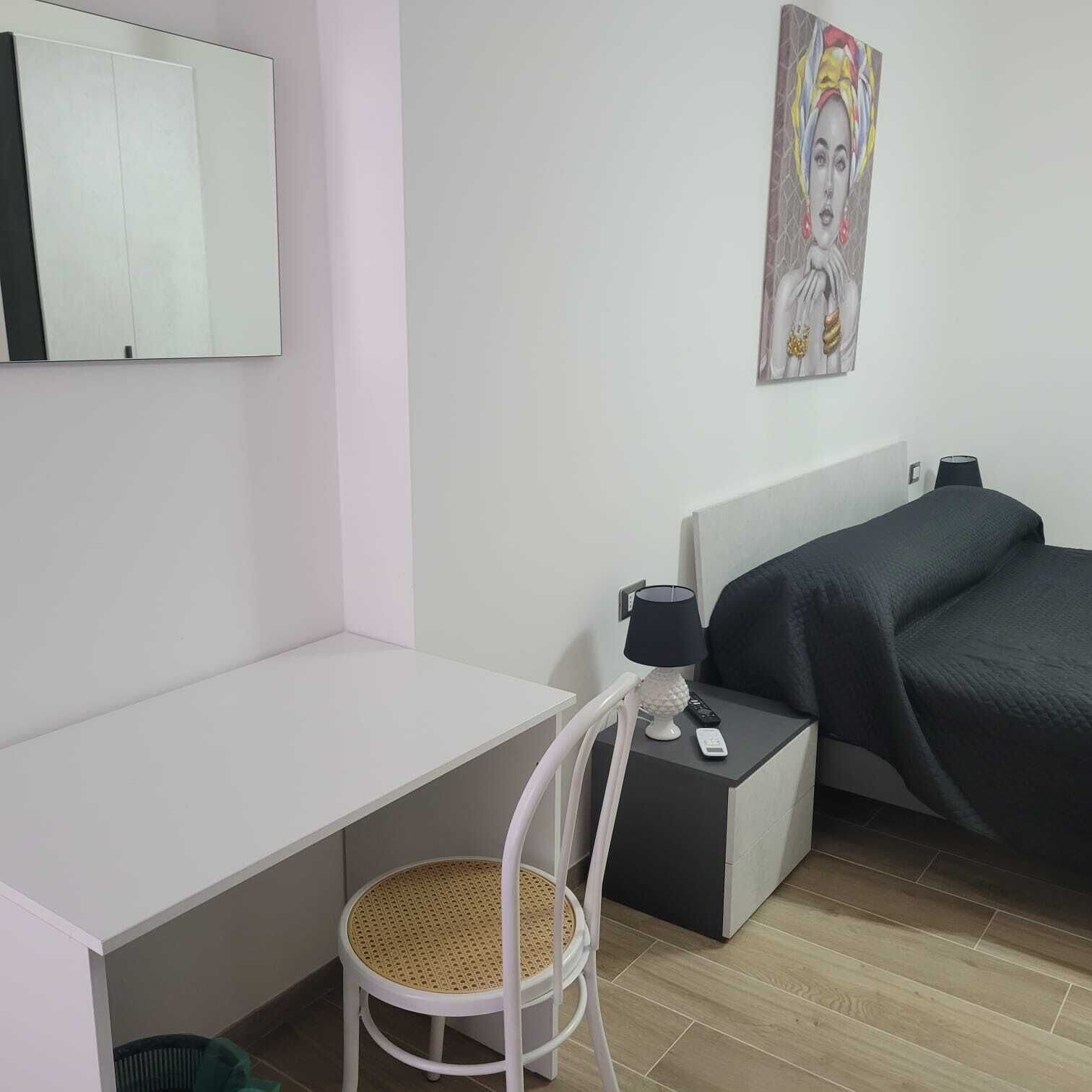 Habitación de hotel con un escritorio blanco, una silla blanca, una cama con cubrecama oscuro, un cuadro en la pared y una lámpara en la mesita de noche.