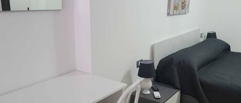 Habitación de hotel con un escritorio blanco, una silla blanca, una cama con cubrecama oscuro, un cuadro en la pared y una lámpara en la mesita de noche.