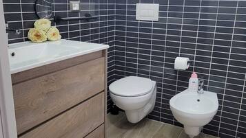 A bathroom with a wooden vanity, a toilet, a bidet, and black tiled walls.