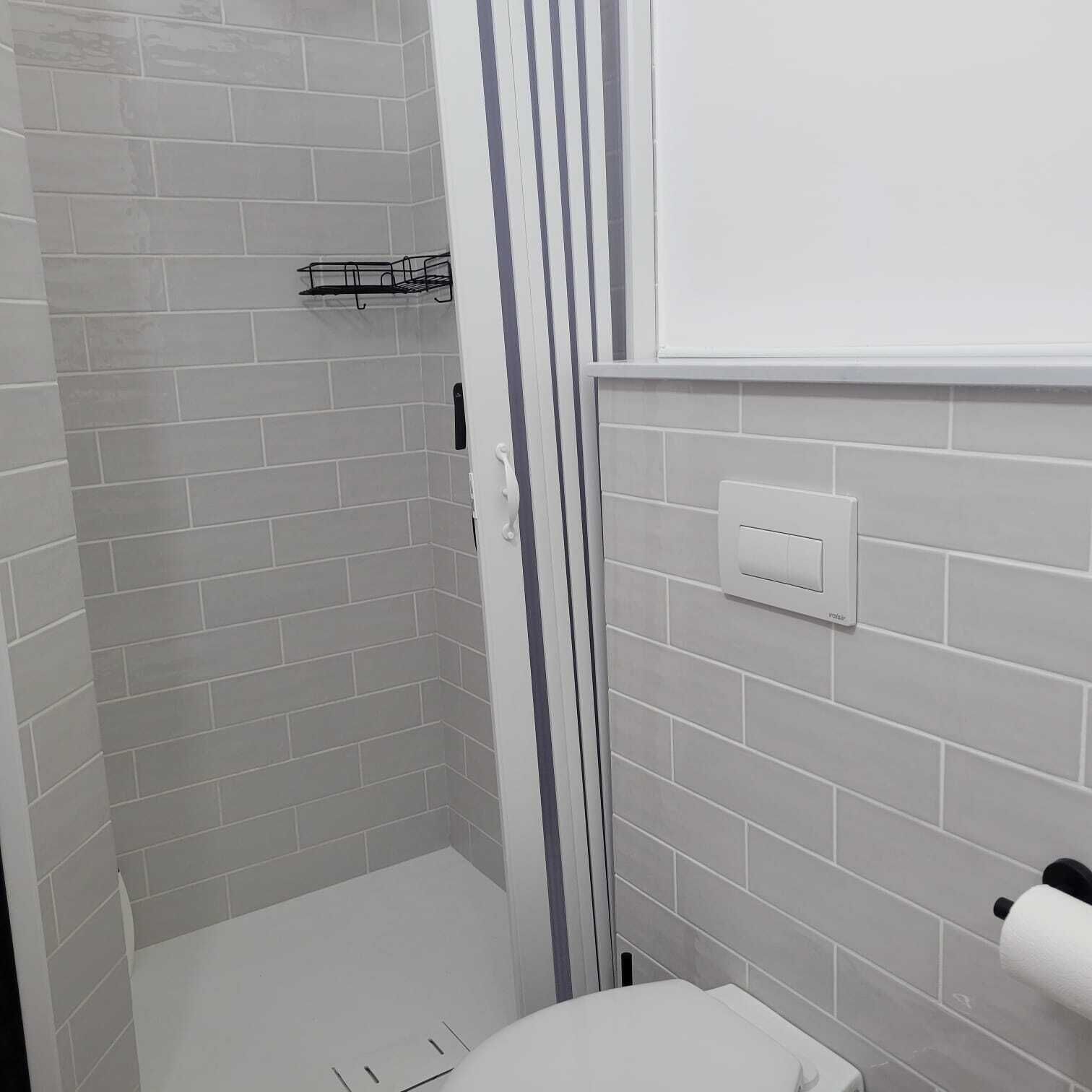 Un baño con azulejos blancos, zona de ducha, inodoro y dispensador de toallas.