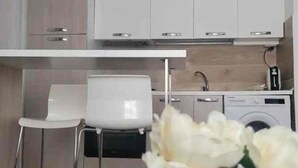 A modern kitchen with white cabinetry, a built-in oven, and a coffee machine. There are two white chairs and a washing machine in the background. - beb dei sogni (Cosenza)