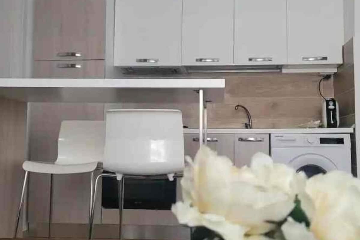 A modern kitchen with white cabinetry, a built-in oven, and a coffee machine. There are two white chairs and a washing machine in the background.