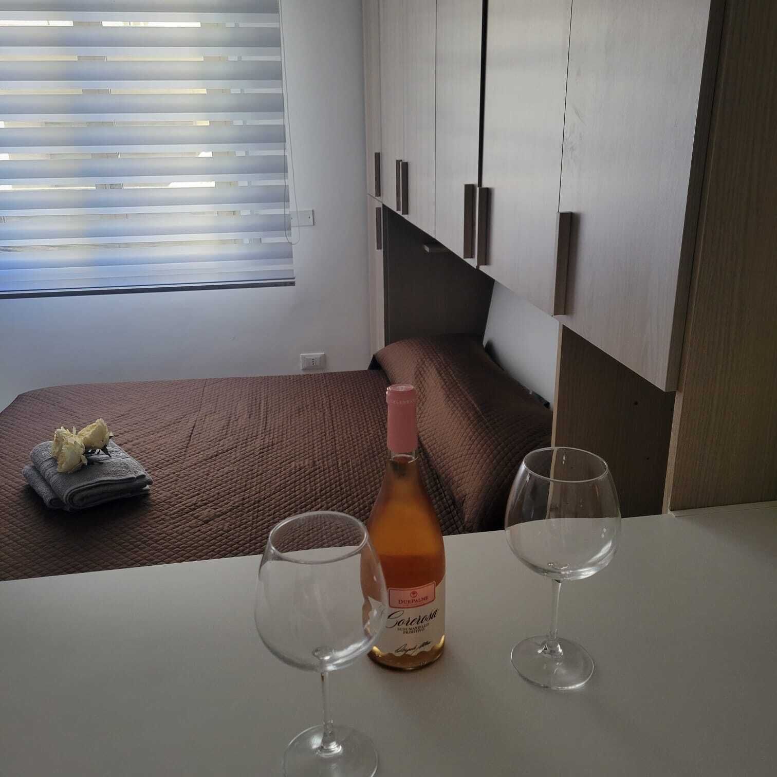 A hotel room with a bed, a table with two wine glasses, a bottle of wine, and a window with blinds.