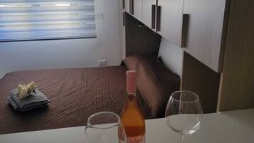 A hotel room with a bed, a table with two wine glasses, a bottle of wine, and a window with blinds.