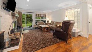A living room with a fireplace, a television mounted on the wall, a sofa, a coffee table, and a large window with a view of the outdoors. - Inviting Ski-In/Ski-Out Condo at Okemo +Hot Tub (Ludlow)