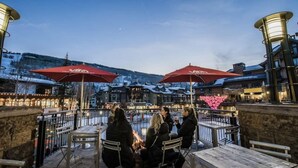 Een terras met tafels en stoelen, rode parasols en uitzicht op een besneeuwd berglandschap.
