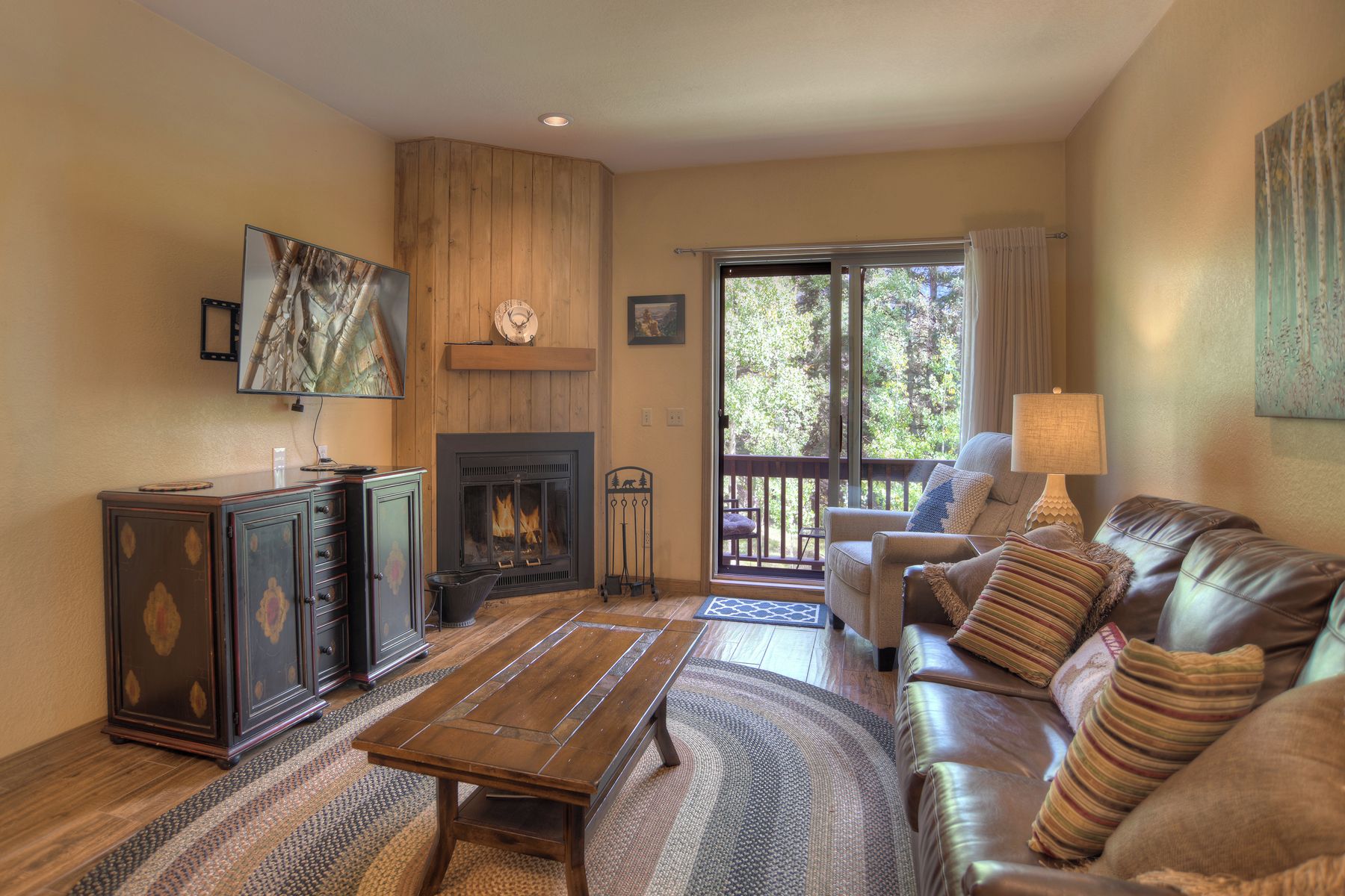 A cozy living room with a fireplace, leather sofa, wooden cabinet, and a large window with a view of trees.