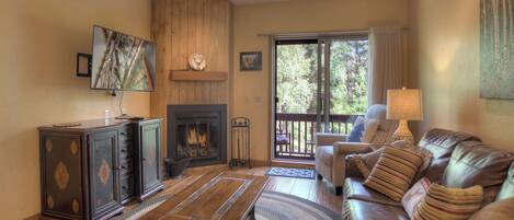 A cozy living room with a fireplace, leather sofa, wooden cabinet, and a large window with a view of trees.