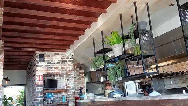 A rustic interior with exposed brick walls, wooden ceiling beams, a brick oven, and a counter with plants. - This is New England! (Armidale)