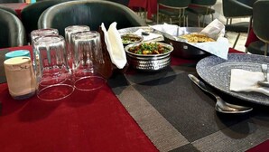 A dining table with a red tablecloth, clear glasses, a metal bowl with food, and a spoon. - Hotel Devine  (Zirakpur)