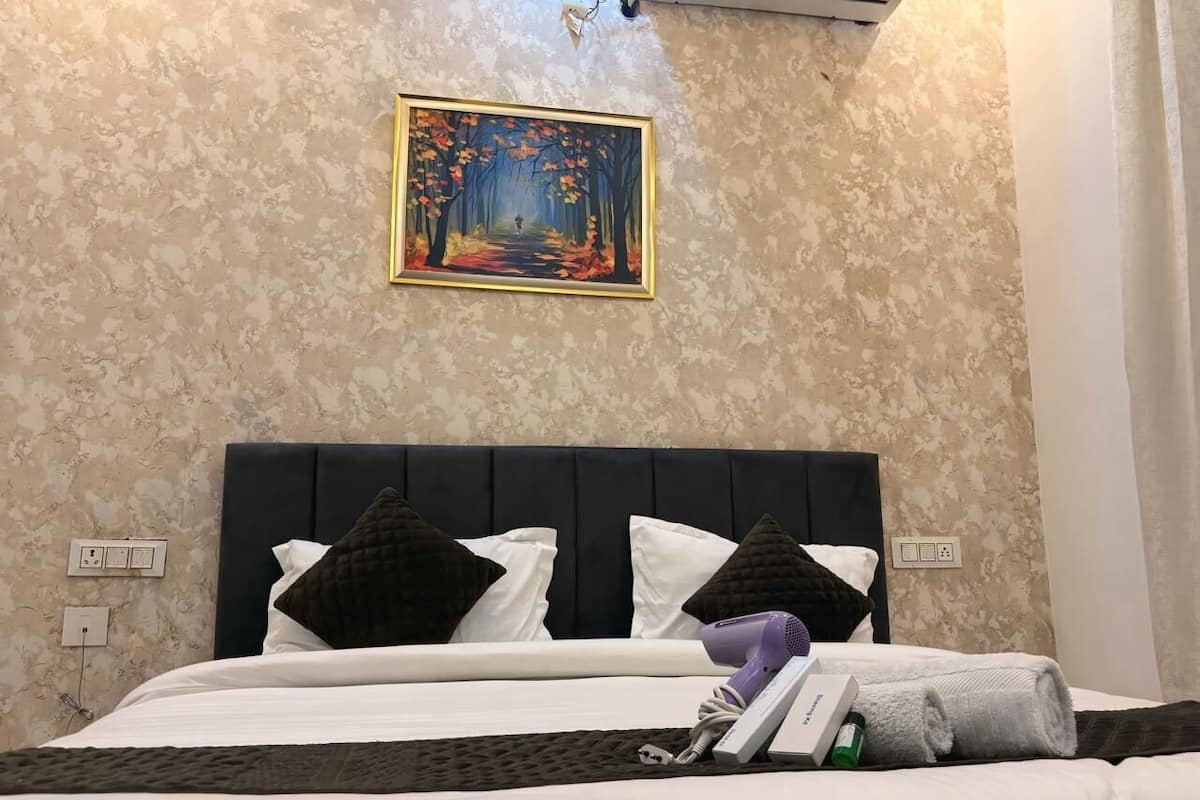 A neatly made bed with white linens and dark pillows, a painting of a forest on the wall, and a power outlet on the wall.