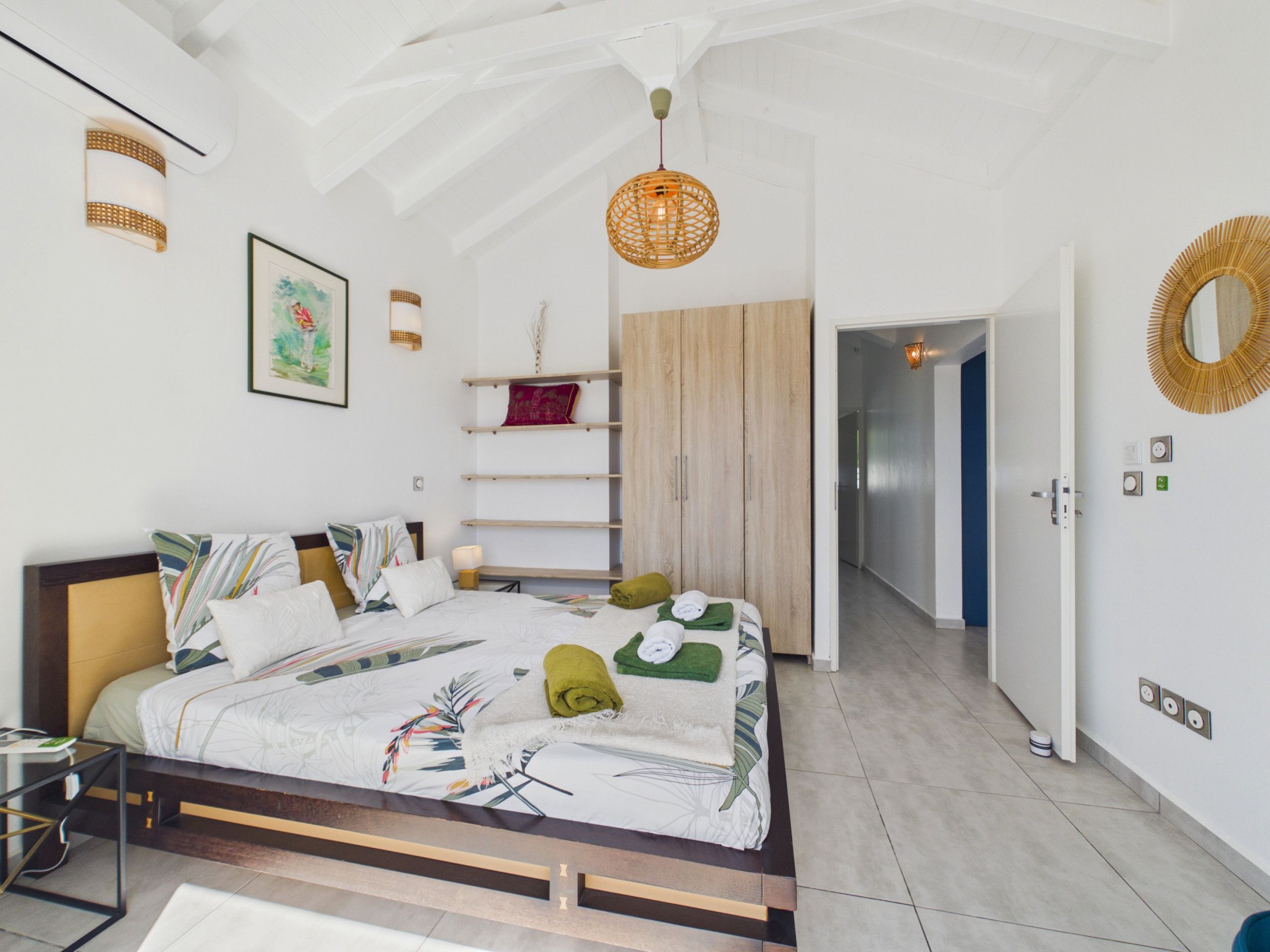 A bedroom with a bed, bedside table, wall-mounted shelf, and a door leading to another room.