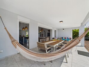 A hammock in a spacious patio area with a dining table and chairs, a kitchen in the background, and a view of the outdoors.