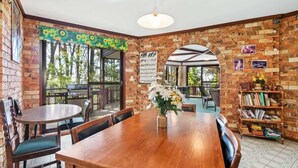 A dining area with a brick wall, a wooden table, chairs, and a bookshelf. - Towering Gums (Lilli Pilli)