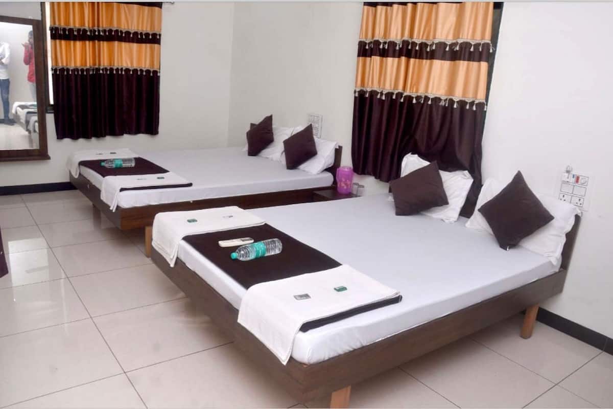 A hotel room with three single beds, each with a white mattress, brown headboard, and a black blanket. There is a small table between the beds with a bottle of water and a green object on it. The room has white walls and a tiled floor.