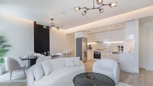 Een moderne woonkamer met een witte bank, een glazen salontafel en een eethoek met een tafel voor vier personen. In de achtergrond is een keuken met witte kasten en roestvrijstalen apparatuur zichtbaar.