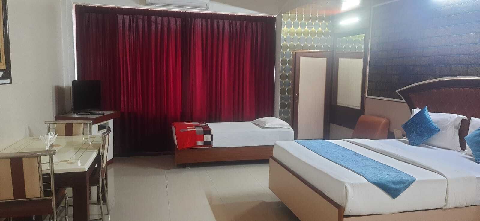 A hotel room with a bed, a desk with a chair, a television, and a window with red curtains.