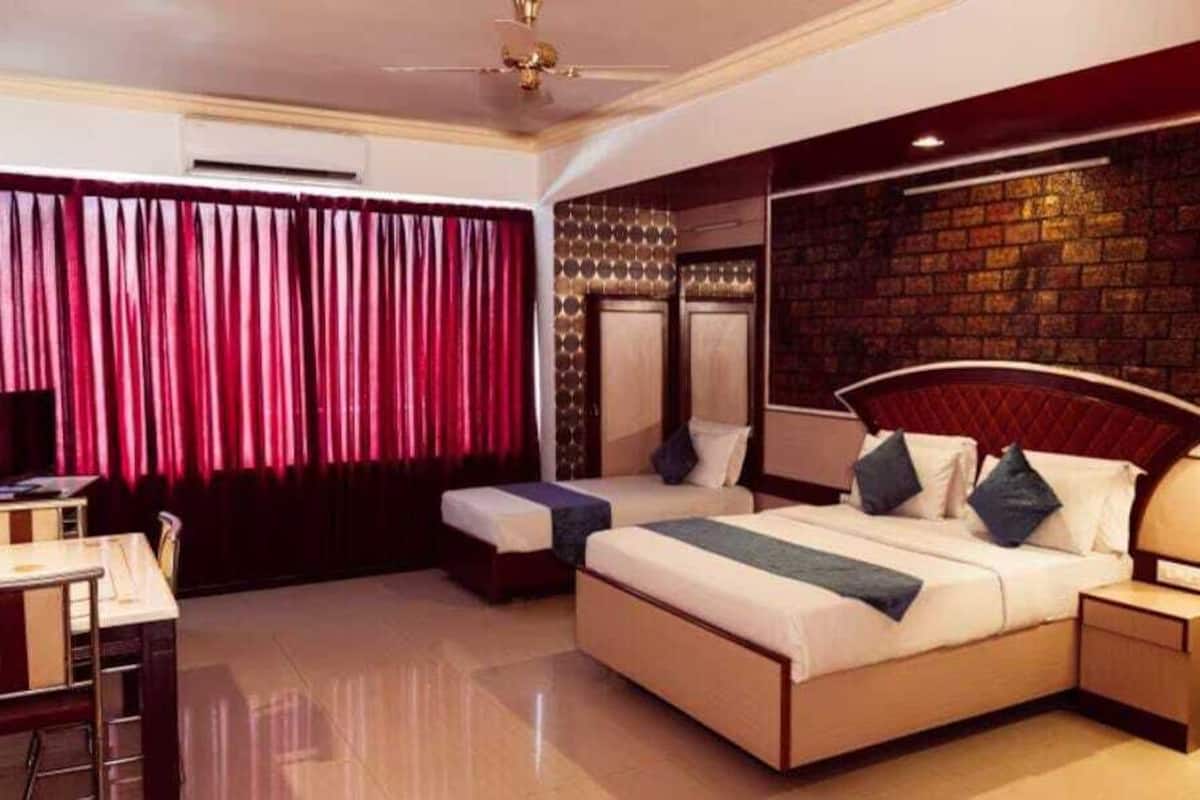 A hotel room with a large bed, a desk, and a chair. There is a window with curtains, a wall with a brick pattern, and a ceiling fan.