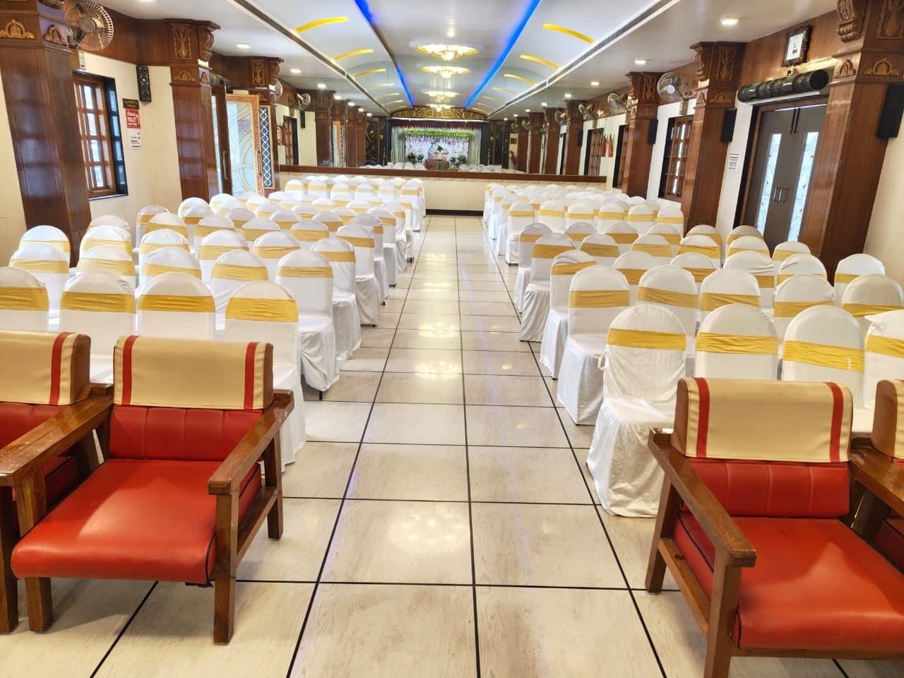 Une salle de banquet avec des rangées de chaises et de tables, une scène à l’avant et des panneaux de bois aux murs.