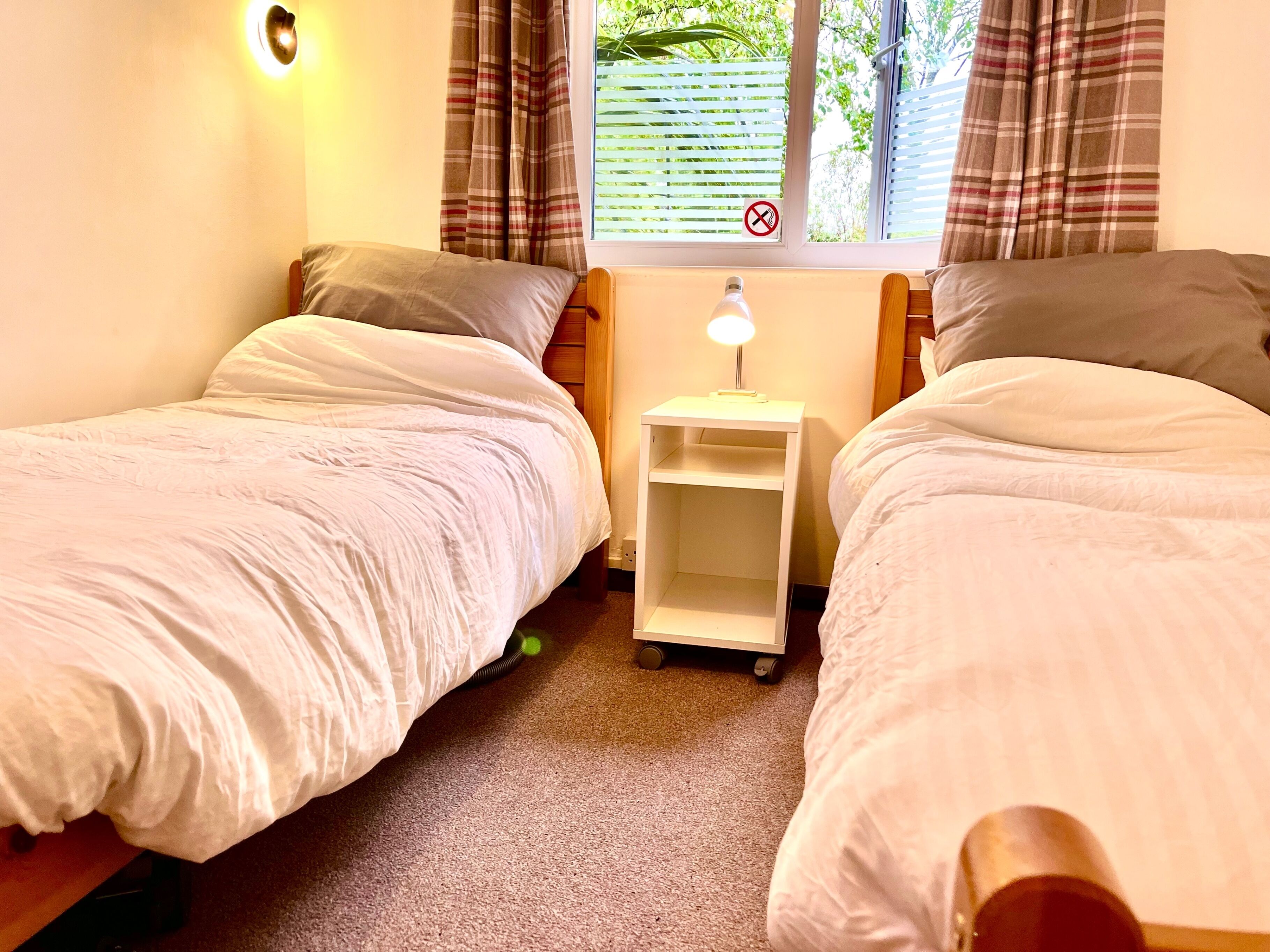 A room with two single beds, a small bedside table with a lamp, a window with blinds, and a wall-mounted light fixture.