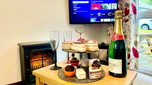 A cozy room with a fireplace, a television displaying a streaming service, and a table with pastries and a bottle of champagne. - 2 Bedrooms Cornish Holidays For Penzance & St Ives (Penzance)