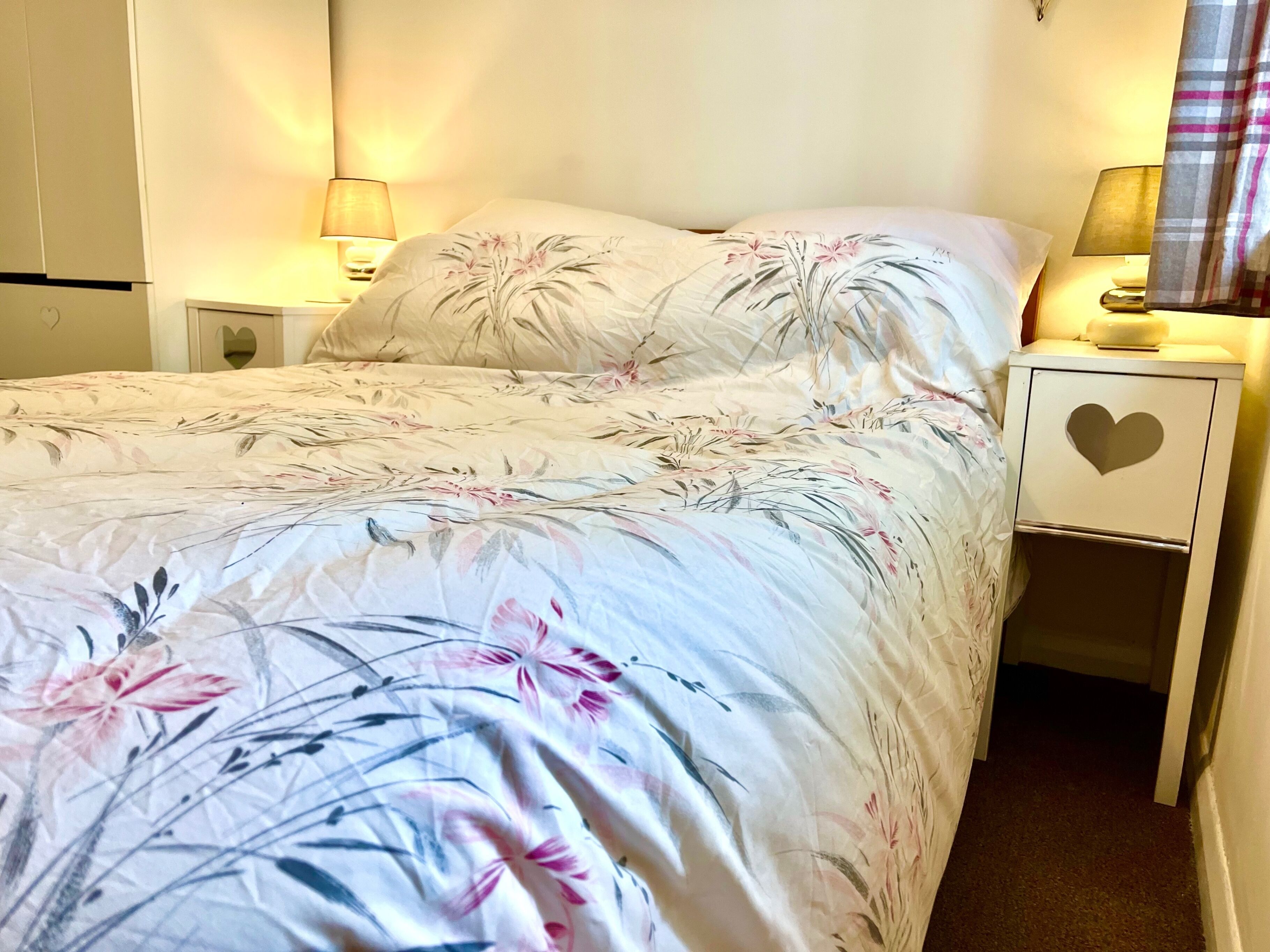 A bed with floral bedding, two bedside lamps, a heart-shaped drawer, and a heart-shaped mirror on the nightstand.