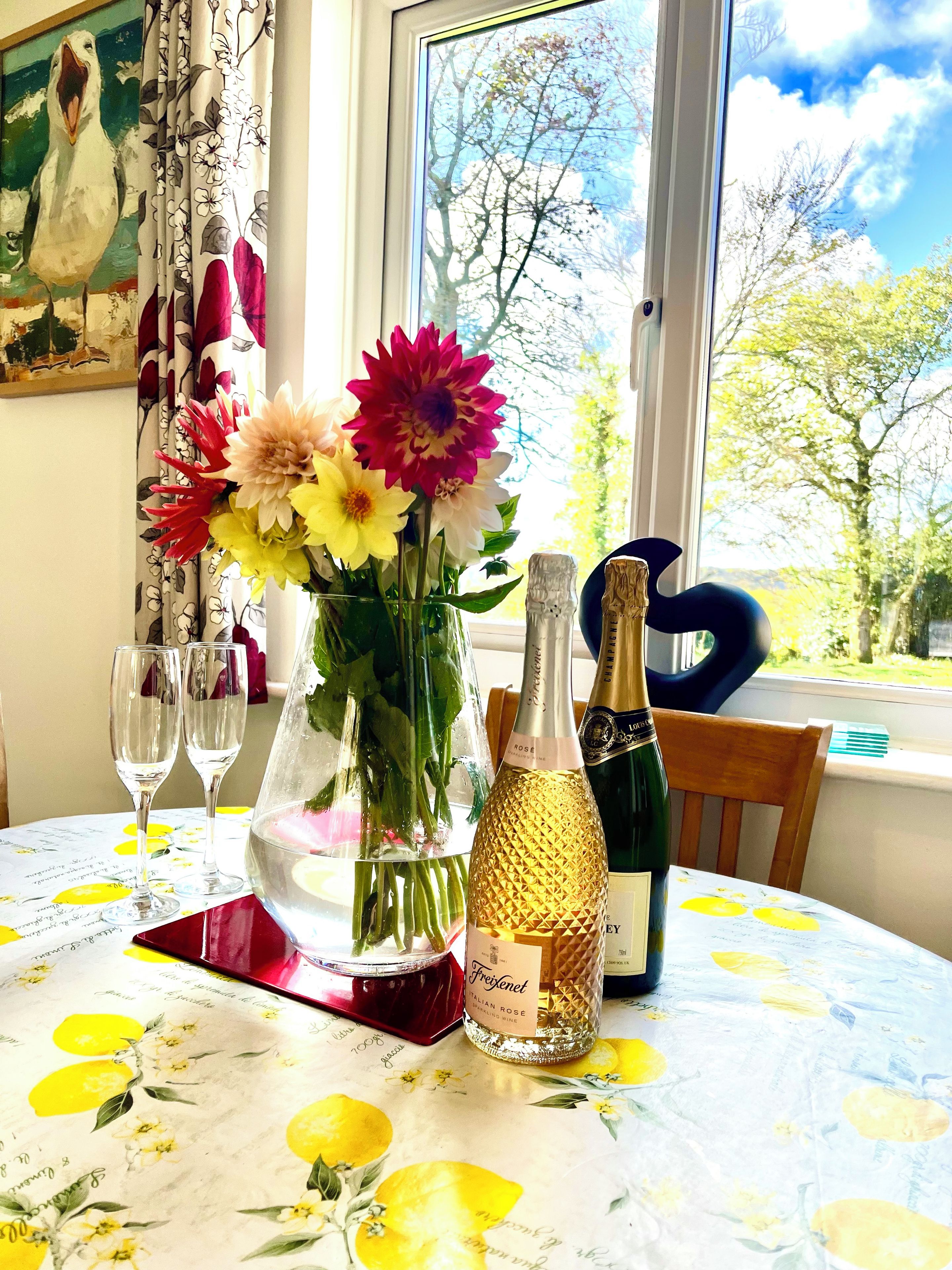 A table with a floral tablecloth, a vase with flowers, two glasses, a bottle of champagne, and a decorative item.