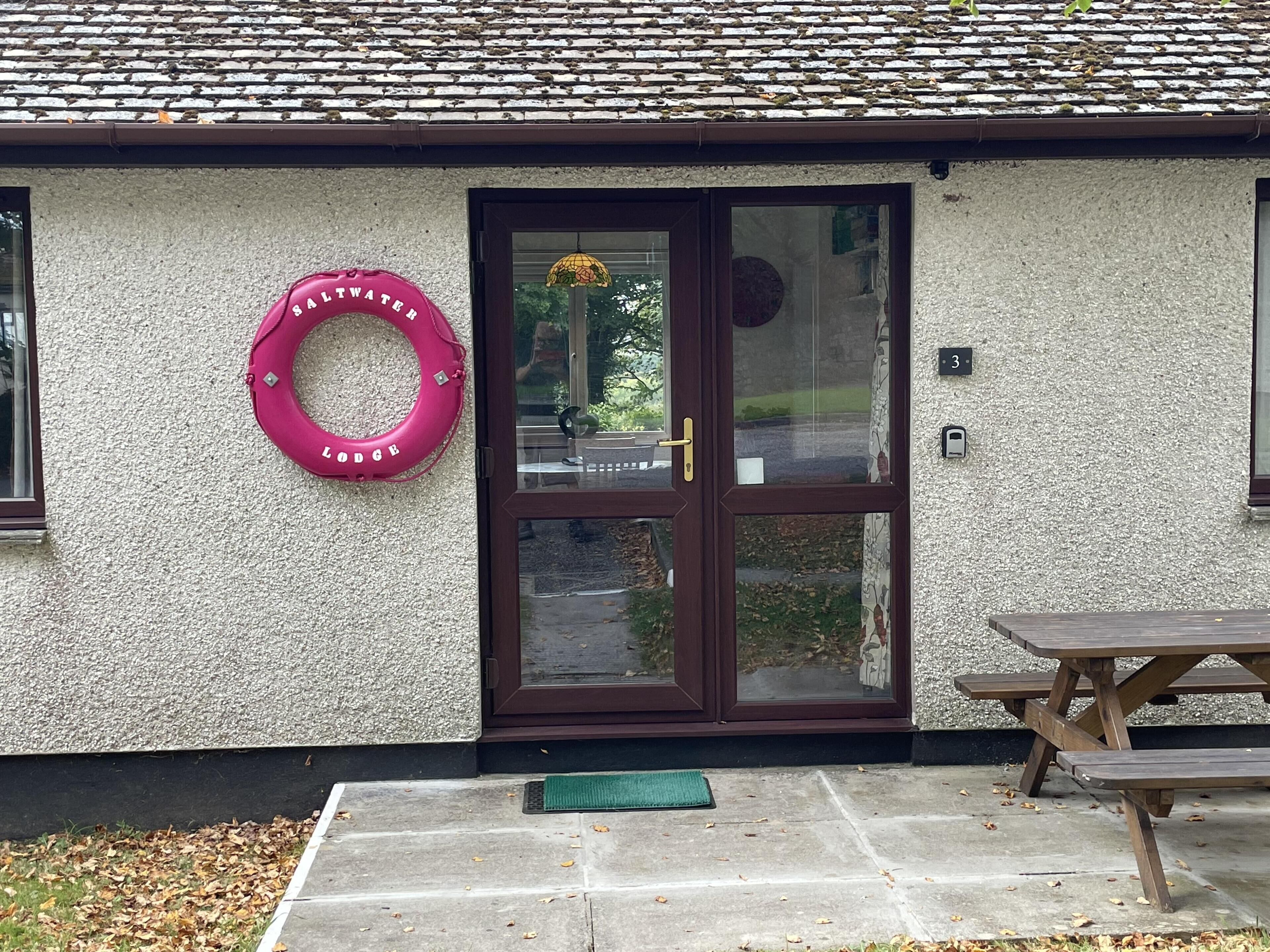 L’entrée du chalet est dotée d’une porte vitrée, d’une bouée de sauvetage rose et d’une table de pique-nique à l’extérieur.