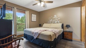 Une chambre à coucher comprenant un lit, un ventilateur de plafond, une table de chevet, une chaise et une fenêtre avec des rideaux.