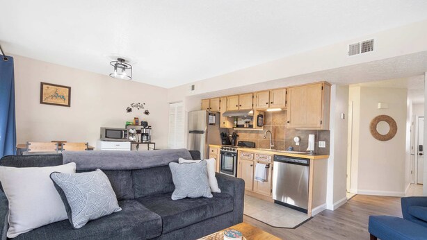 A modern living room with a grey sofa, a coffee table, and a kitchen area with wooden cabinets and stainless steel appliances. - The Nautical Nook – Charming Walk-In Condo for 4! (Branson)