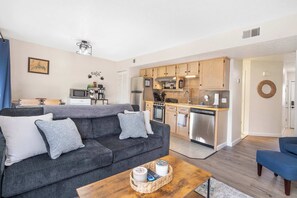 A modern living room with a grey sofa, a coffee table, and a kitchen area with wooden cabinets and stainless steel appliances. - The Nautical Nook – Charming Walk-In Condo for 4! (Branson)