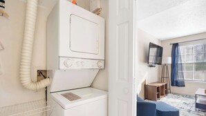 A laundry room with a white washer and dryer, a wooden shelf, and a blue chair. - The Nautical Nook – Charming Walk-In Condo for 4! (Branson)