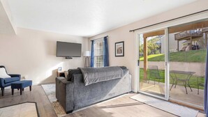 A living room with a grey sofa, a TV, and a sliding glass door leading to an outdoor patio. - The Nautical Nook – Charming Walk-In Condo for 4! (Branson)