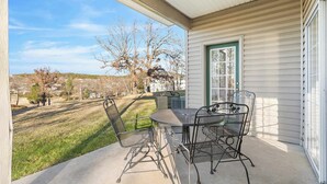 Eine überdachte Terrasse mit einem Metalltisch und -stühlen, eine Tür mit grünem Rahmen und Blick auf einen Rasen und Bäume.