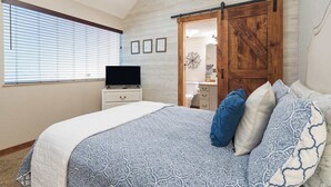 Een slaapkamer met een groot bed, een televisie op een dressoir, een schuifdeur in boerderijstijl en een raam met jaloezieën.
