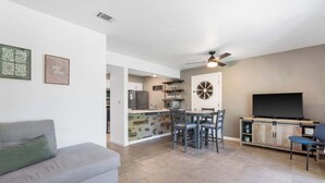 A modern living room with a gray sofa, a dining area with a table and chairs, a kitchen area with a refrigerator and cabinets, and a flat-screen TV on a wooden cabinet.