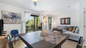 A modern living room with a large table, a sofa, and a TV. There are curtains, a painting, and a clock on the wall.