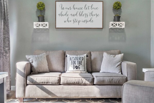 A neutral-toned sofa with a cushion that reads "RESERVED FOR THE DOG" and a framed quote on the wall above.