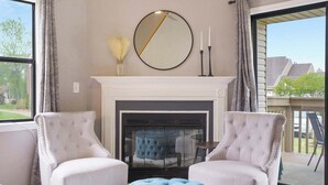 Un salon avec une cheminée, deux fauteuils, un miroir rond et une vue sur l’extérieur.