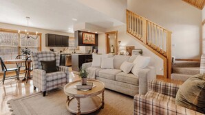 A spacious living room with a sofa, armchairs, and a coffee table. A kitchen is visible in the background, and a staircase leads to an upper level.