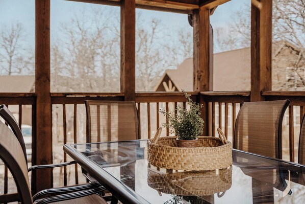 Una terraza de madera al aire libre con una mesa y sillas de vidrio, una cesta tejida con una planta, y vistas a árboles y edificios.
