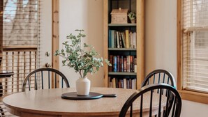 Une table de salle à manger ronde en bois, sur laquelle est posé un vase blanc contenant des plantes vertes, ainsi que des chaises noires et un set de table noir. En arrière-plan, on aperçoit une bibliothèque en bois remplie de livres et un petit modèle de maison.
